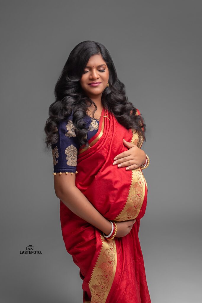 A maternity portrait of a woman in a traditional red sari, with her eyes closed and hands gently cradling her baby bump, captured by Viktoria ILJIN during a professional photoshoot.