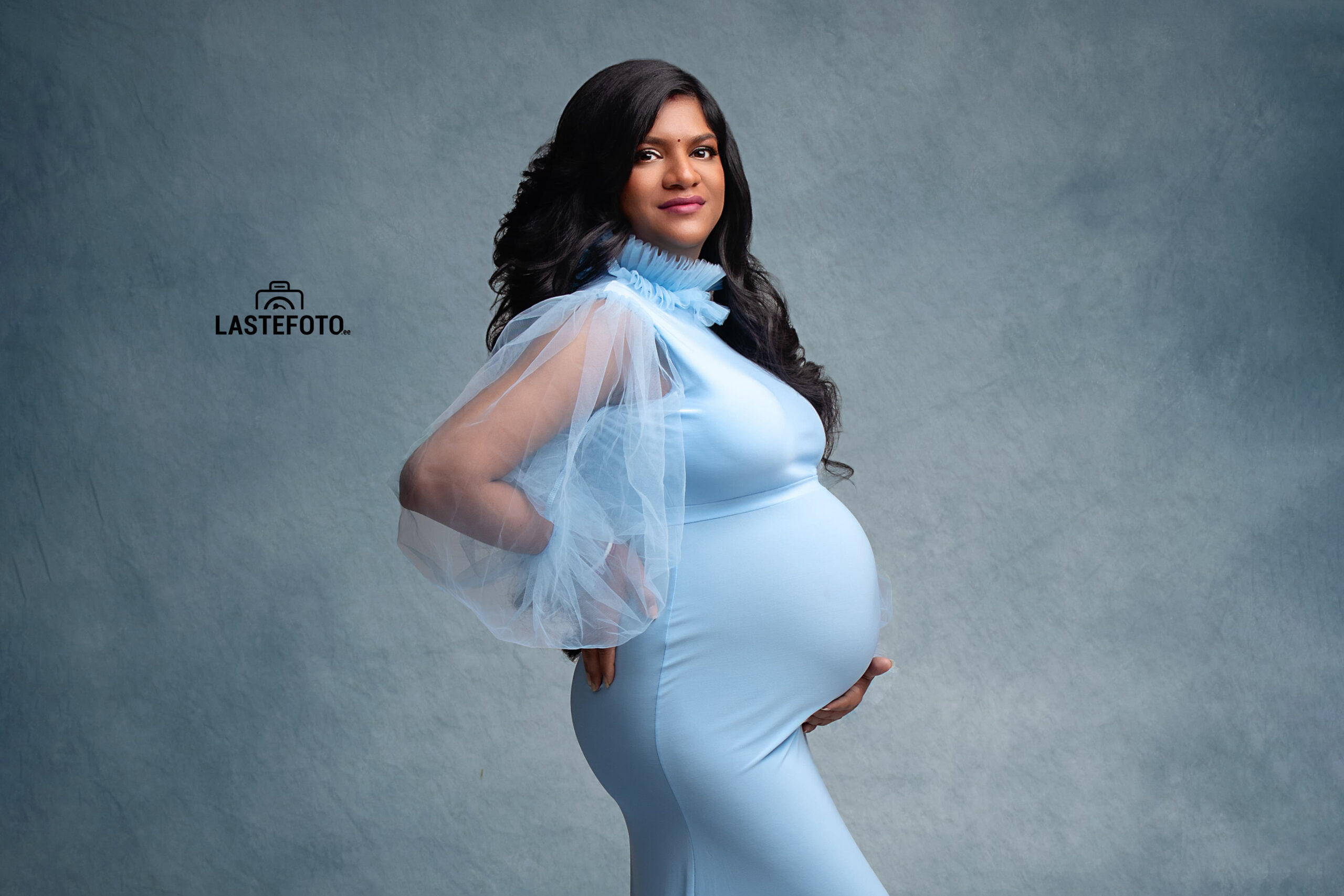 A stunning maternity portrait of a woman in a light blue gown, confidently posing with her hand on her hip, captured by Viktoria ILJIN during a professional photoshoot.