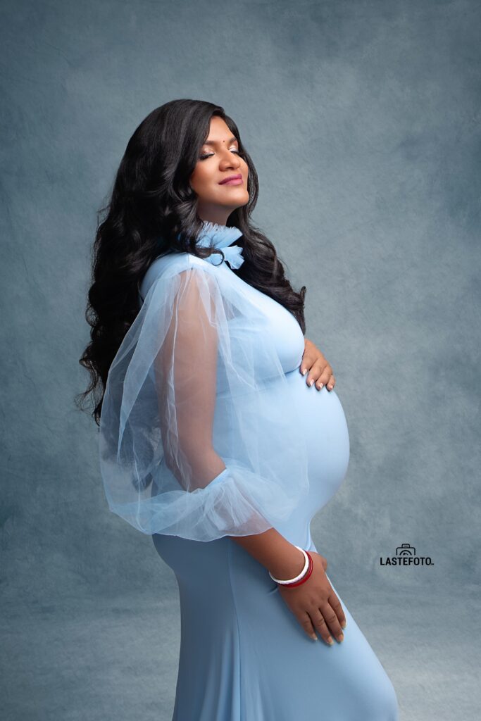 A serene maternity portrait of a woman in a flowing blue gown, eyes closed, with a peaceful expression, captured by Viktoria ILJIN during a professional photoshoot.