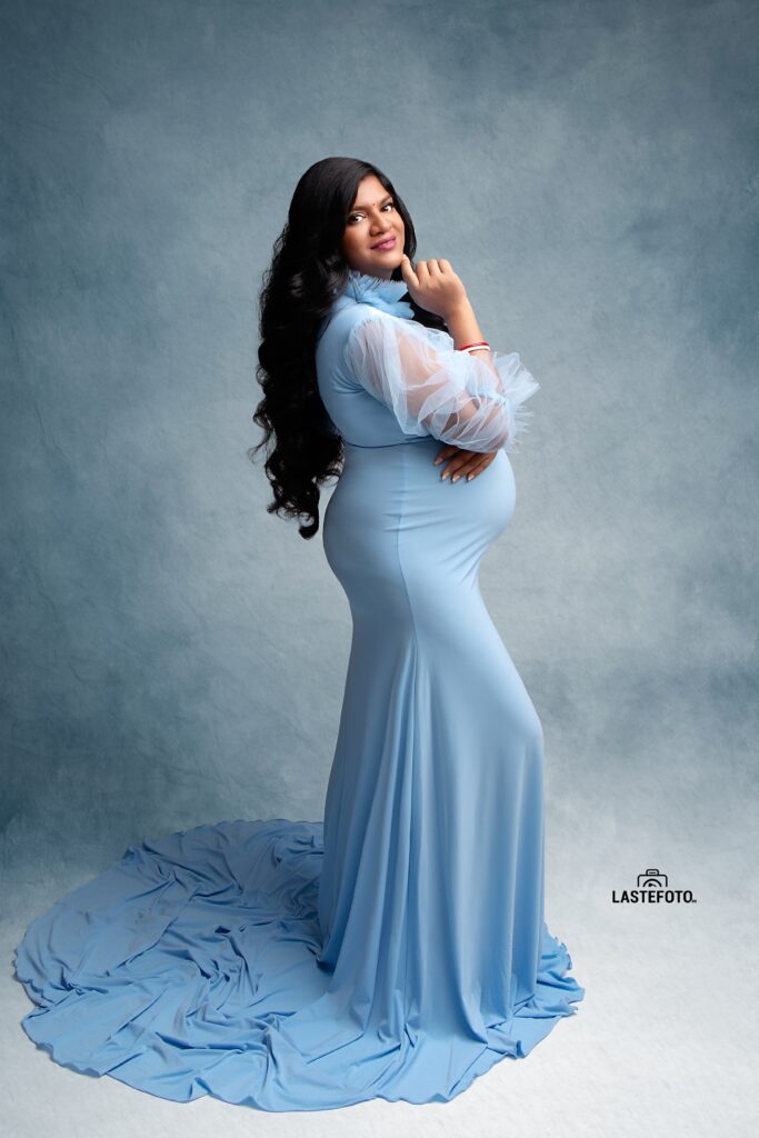 A graceful maternity portrait of a woman in a light blue gown, posing elegantly with her hand resting on her chin, captured by Viktoria ILJIN during a professional photoshoot.