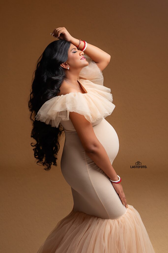 A radiant maternity portrait of a woman in a beige gown, posing with her hand on her head and a serene expression, captured by Viktoria ILJIN during a professional photoshoot.