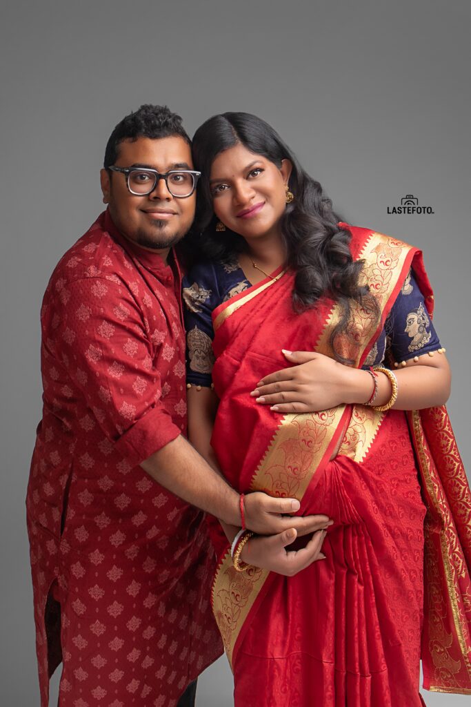 A couple dressed in traditional attire, with the woman wearing a red sari, embracing each other lovingly during a maternity photoshoot, captured by Viktoria ILJIN.