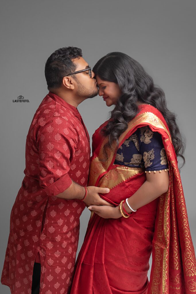 An intimate maternity photo of a couple in traditional attire, with the partner gently kissing the woman's forehead as they hold her baby bump, captured by Viktoria ILJIN.