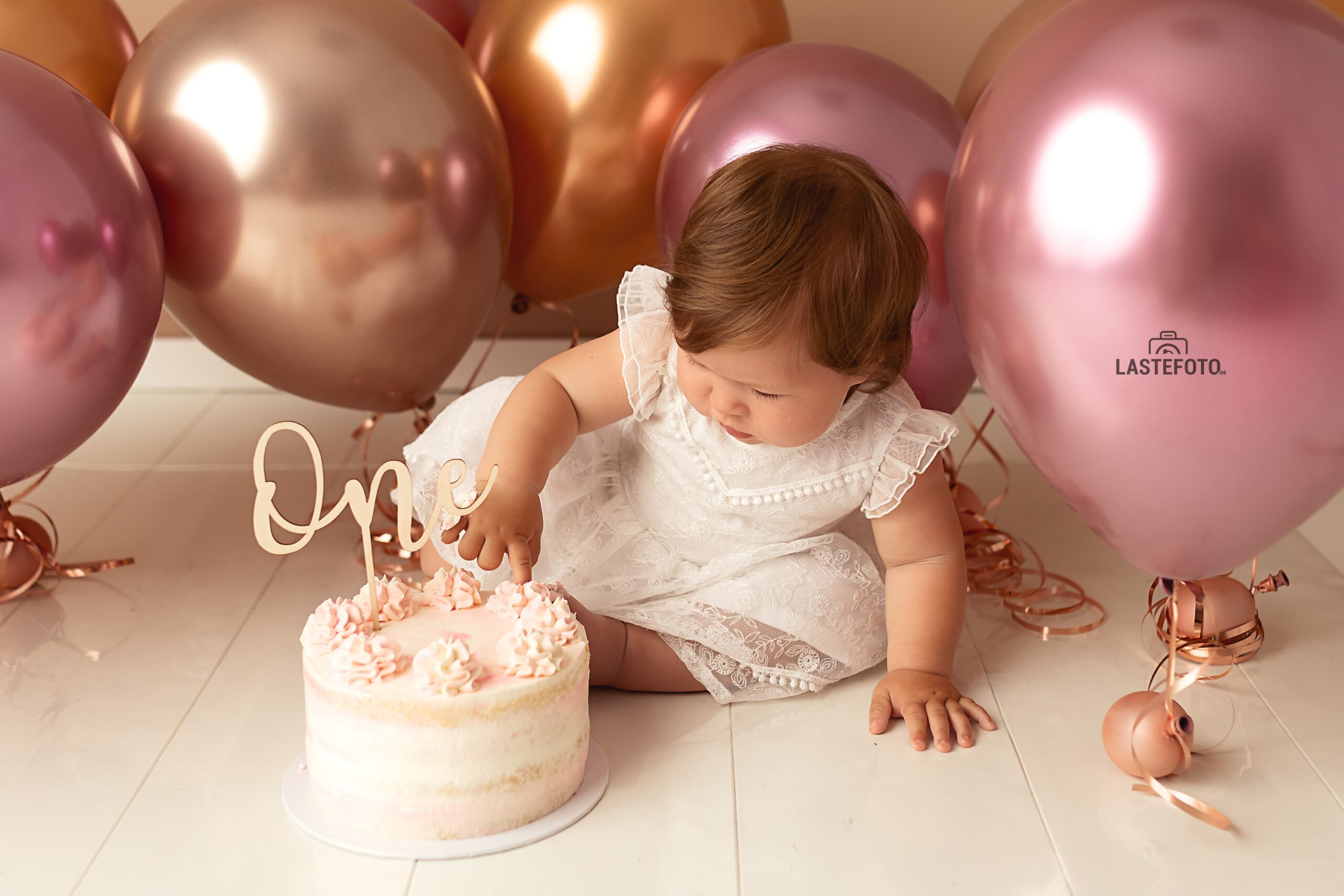 A baby in a white lace dress gently exploring a birthday cake with a "One" topper during a Cake Smash session, surrounded by pink and gold balloons.