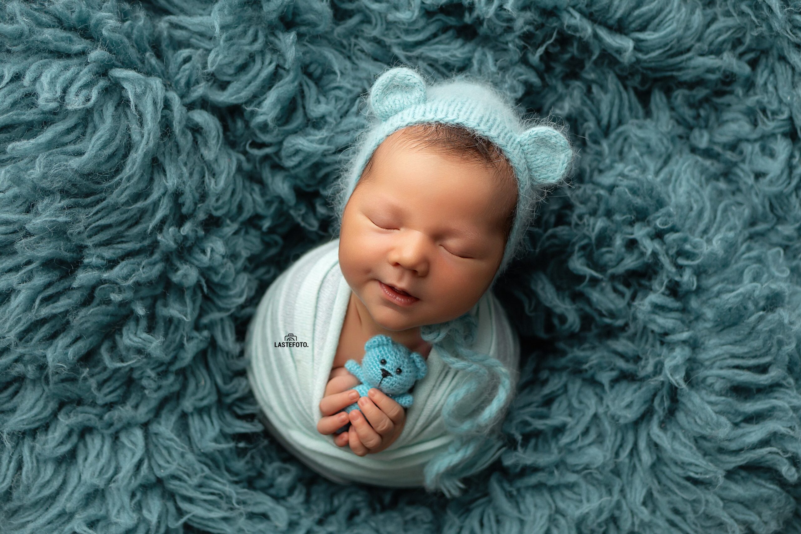 Newborn baby peacefully sleeping on a teal furry blanket, wearing a soft knit hat with bear ears and holding a small knitted teddy bear.