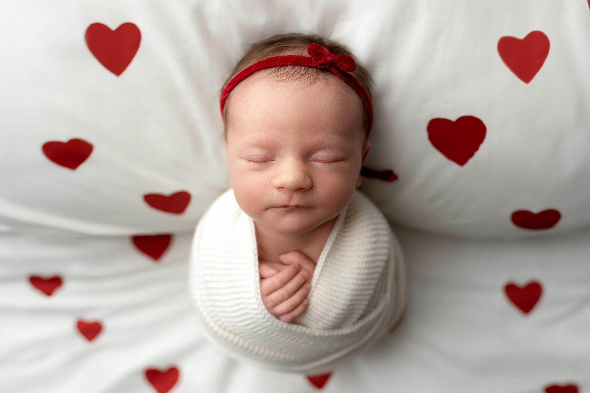 Sleeping newborn wrapped in a white blanket, wearing a red headband, on a heart-themed background.