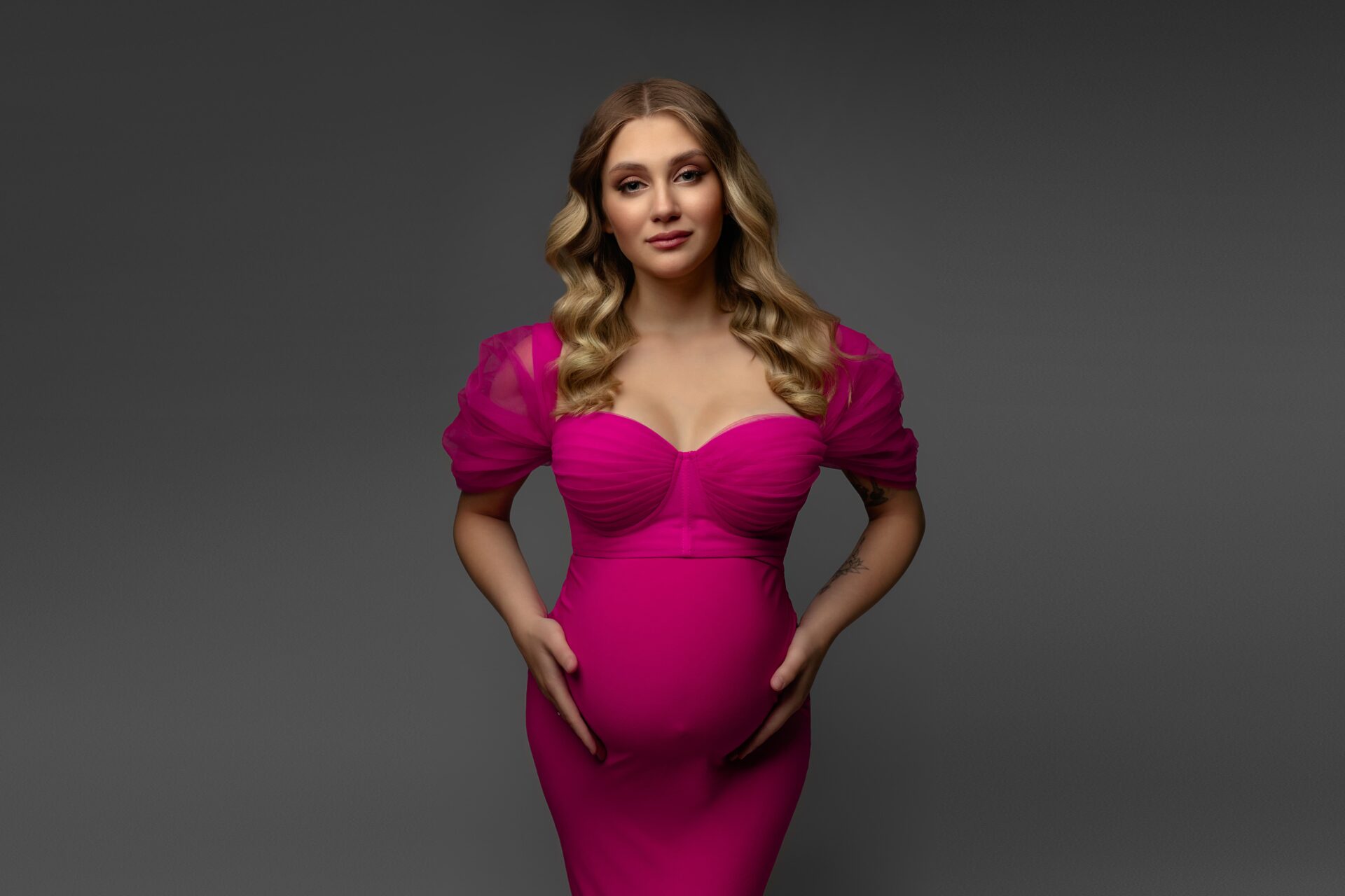 Maternity photoshoot in the studio, woman in a fuchsia dress against a gray background.