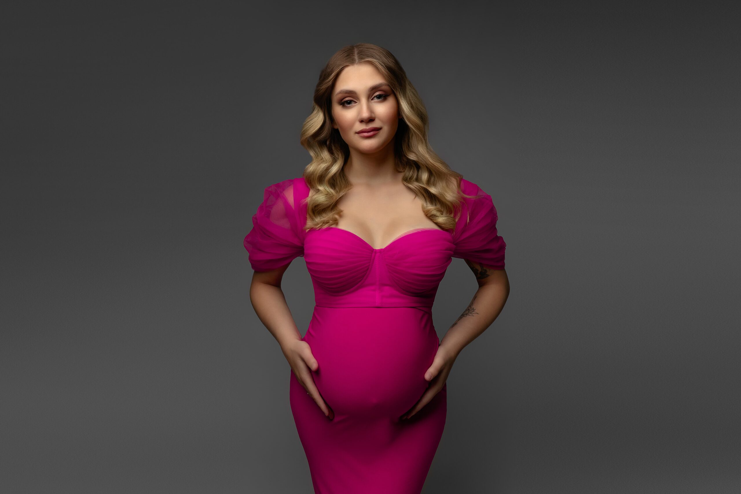 Maternity photoshoot in the studio, woman in a fuchsia dress against a gray background.